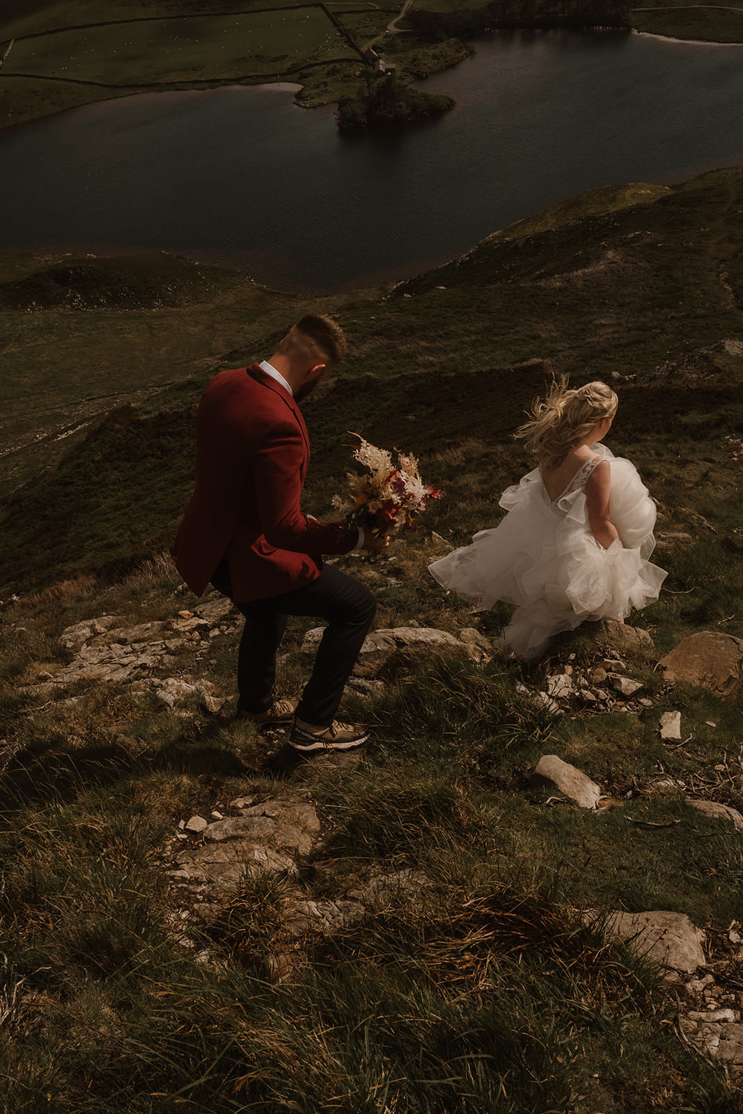 Snowdonia Elopements | bride holding up a big white wedding dress and groom in a red blazer holding a bouquet walking down a mountain near Cadair Idris