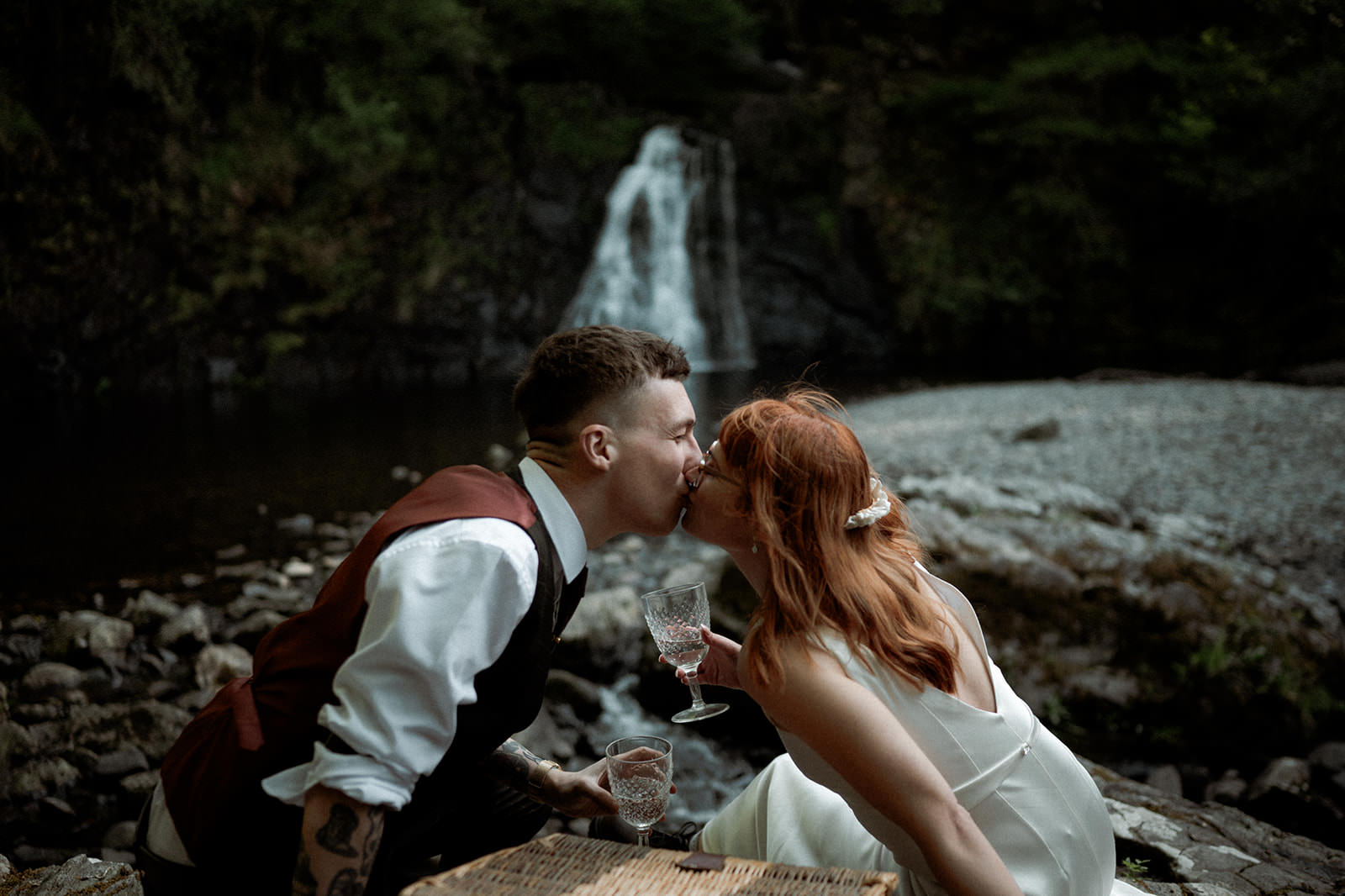 Snowdonia Elopements | bride wearing a wedding dress and groom sharing a kiss while drinking champagne with a waterfall in the background