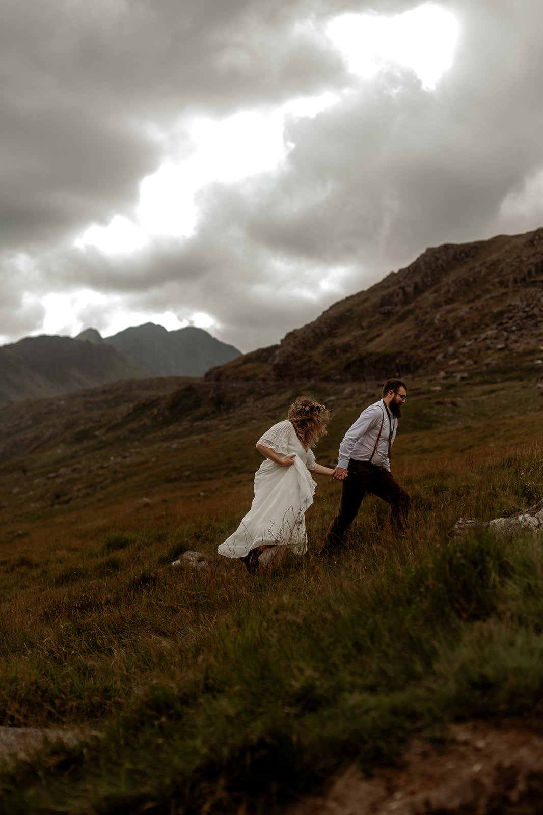 Snowdonia Elopements | bride wearing white boho style wedding dress and groom wearing braces over his shirt walking up a mountain in Wales on their wedding day